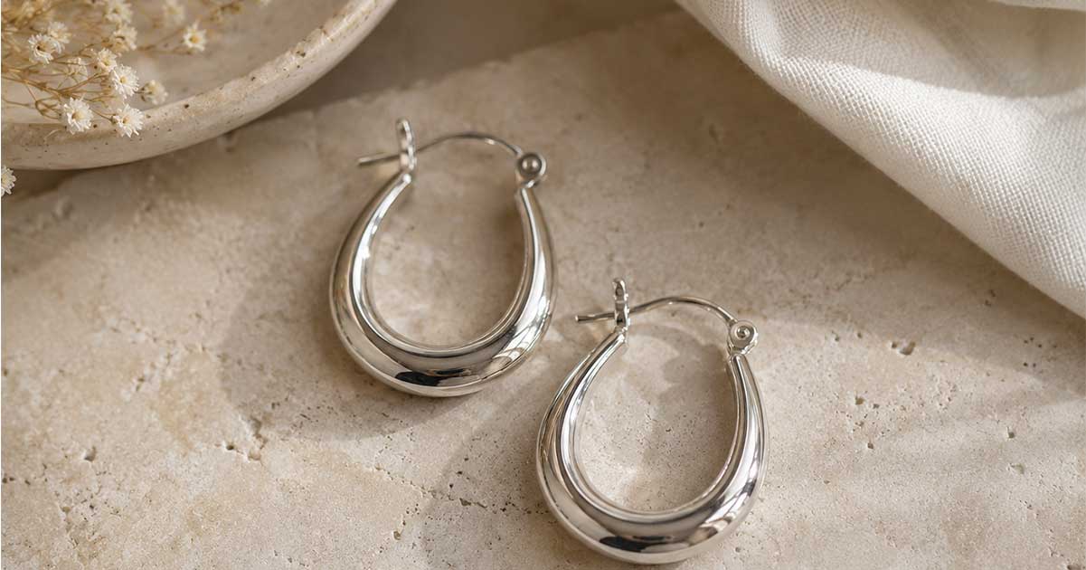 Two oval-shaped silver hoop earrings side-by-side on a beige stone table.