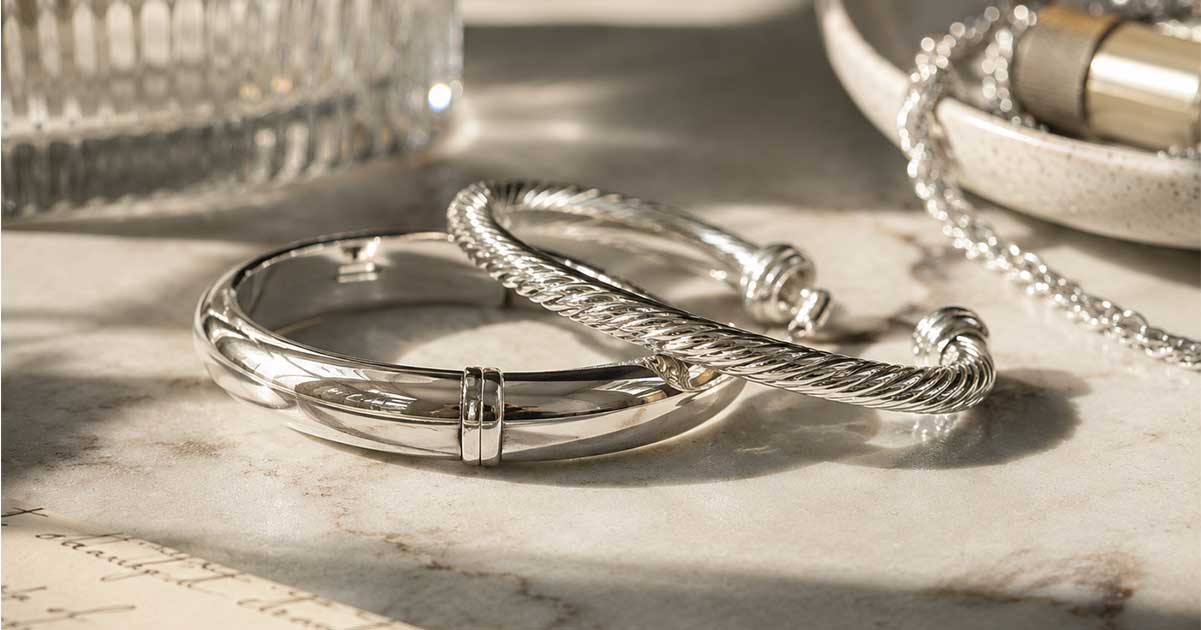 Two silver bracelets, one smooth and the other braided, sitting on a white marble tabletop.
